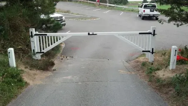 A white, gated barrier blocks a paved driveway leading to a parking area with a vehicle parked in the distance.