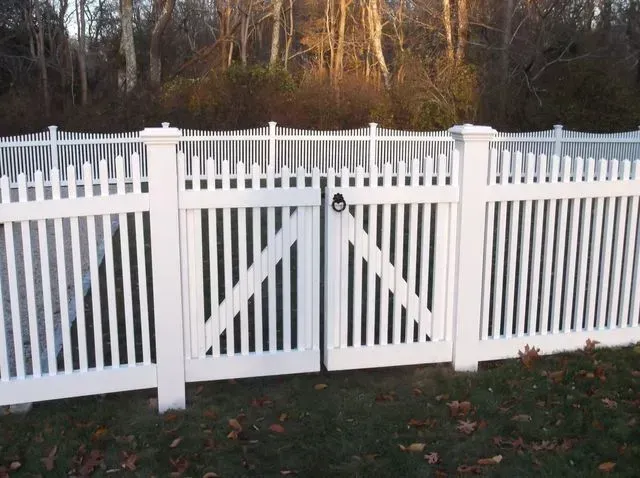 A white vinyl picket fence with a double gate in a grassy backyard setting with trees in the background.
