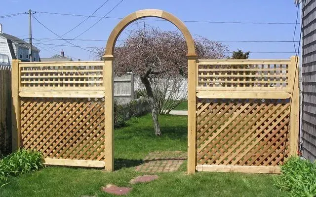 A wooden garden fence featuring a central arched gate, lattice panels, and a view into a backyard with a small tree.