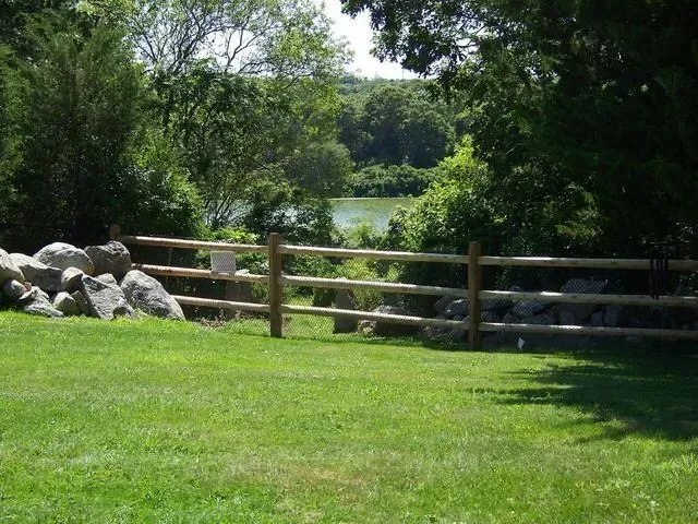 A wooden three-rail fence sits on a green lawn, bordering a pile of rocks and trees overlooking a river.