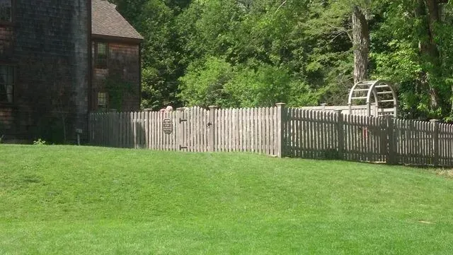 A wooden fence with a gate borders a grassy backyard, situated next to a house with a shingled exterior and a garden arbor.