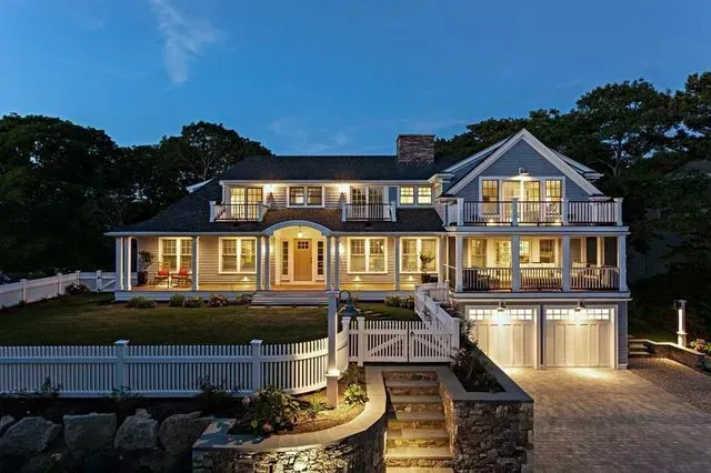 A multi-story gray shingled coastal house at dusk with a white picket fence, lit windows, and a paved driveway.