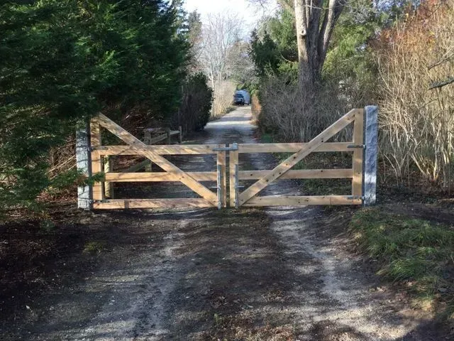 A double wooden farm gate stands closed across a dirt driveway, flanked by stone posts and lush foliage.