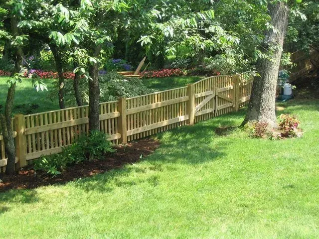 A light-colored wooden picket fence curves through a lush green backyard, lined with mature trees and garden beds.