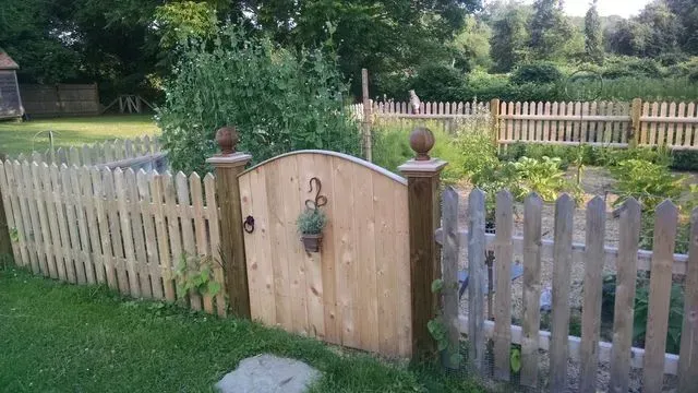 A wooden gate with a hanging basket, set in a picket fence enclosing a garden area with lush plants.