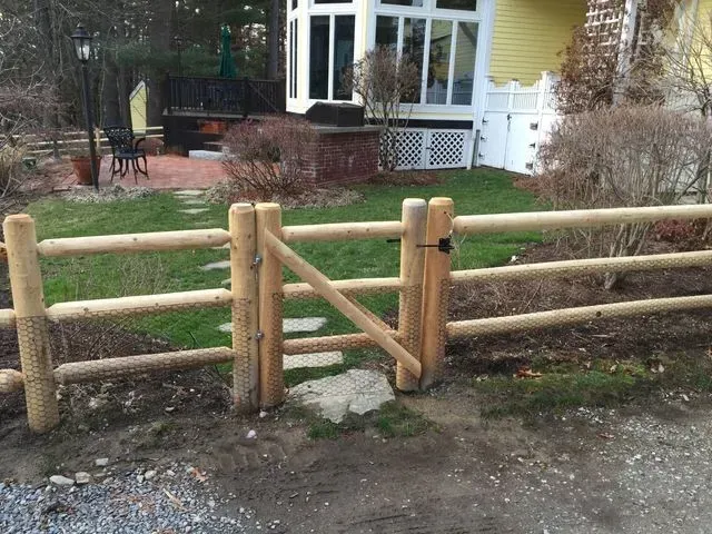 A three-rail wooden fence with a matching gate sits in front of a house with a yellow wall and white window trim.
