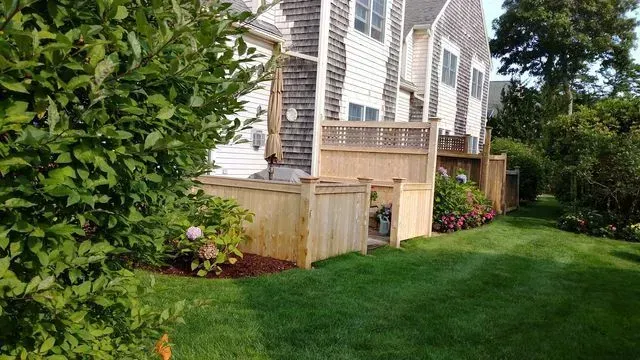 A wooden fence surrounds a patio area in front of a shingled multi-story home, bordered by a lush green lawn and bushes.