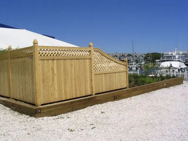 A wooden privacy fence with lattice trim borders a landscaped bed set in white gravel near a boat marina.