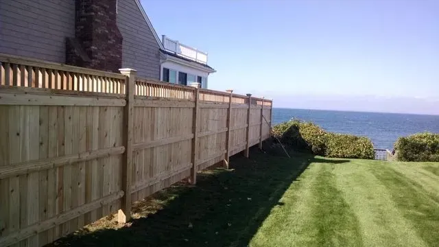 A wooden fence stretches along a grassy lawn toward the ocean, situated next to the side of a house.