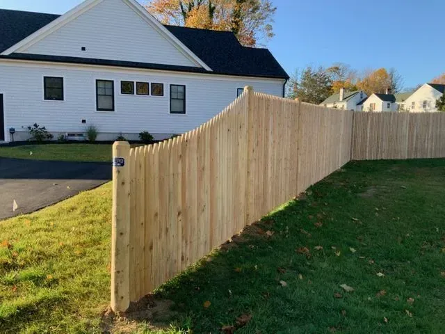 A curved wooden picket fence stands in a grassy yard in front of a white house on a sunny day.
