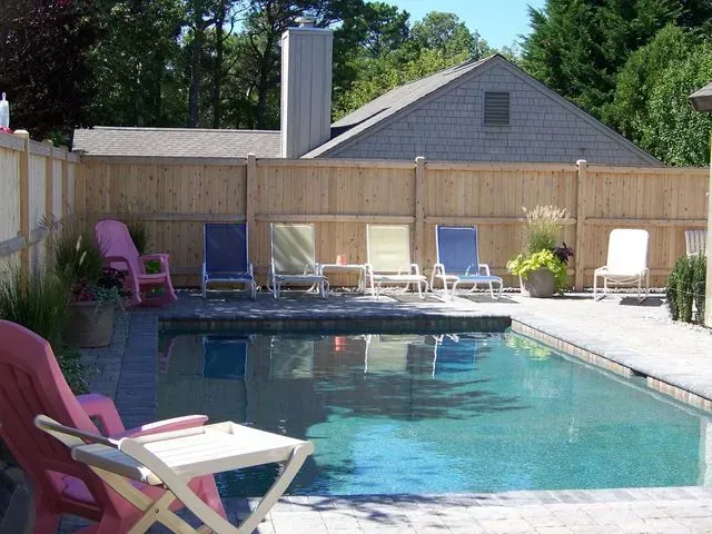A backyard swimming pool with a row of lounge chairs on a patio, enclosed by a wood fence with a house in the background.