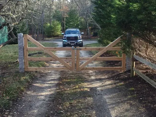 A closed wooden gate across a gravel driveway, with a parked truck visible in the background among trees.