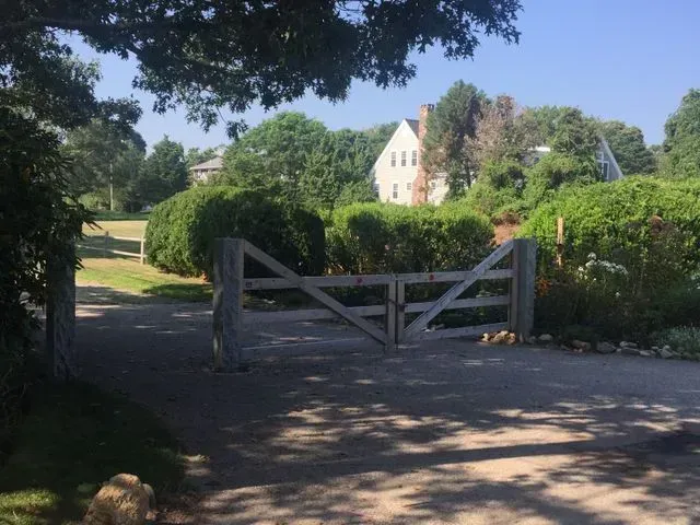 A wooden gate across a gravel driveway, leading to a white house surrounded by green trees and bushes under a blue sky.