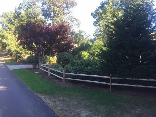 A wooden split-rail fence runs along a paved path next to a lush garden with green shrubs and a dark-leafed tree.