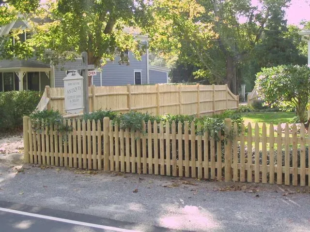 A wooden picket fence sits in the foreground with a taller, solid wood privacy fence behind it in a sunny, tree-lined yard.
