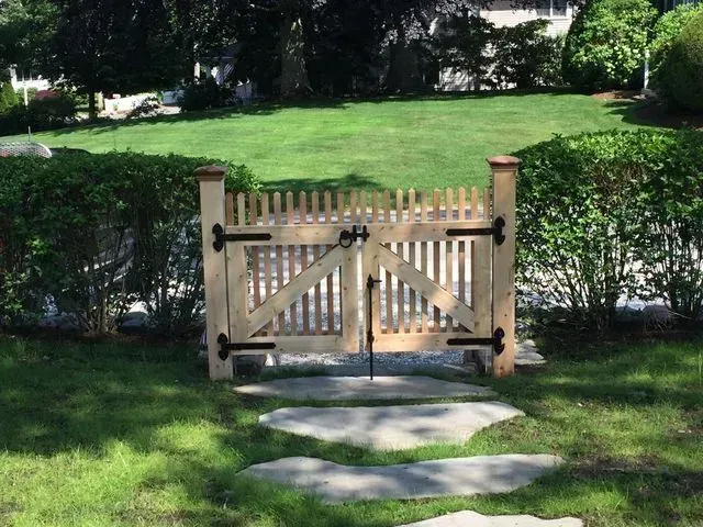 A light-colored wooden double gate with black iron hinges set in a green garden between two hedge rows.