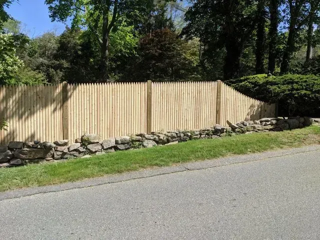 A new wood picket fence sits behind a stone retaining wall along a residential road with green trees in the background.