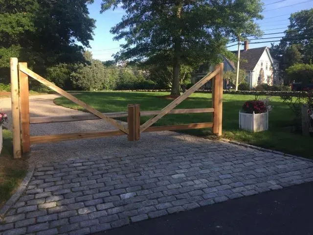 A wooden split-rail driveway gate centered on a cobblestone entrance, with a green lawn and house in the background.