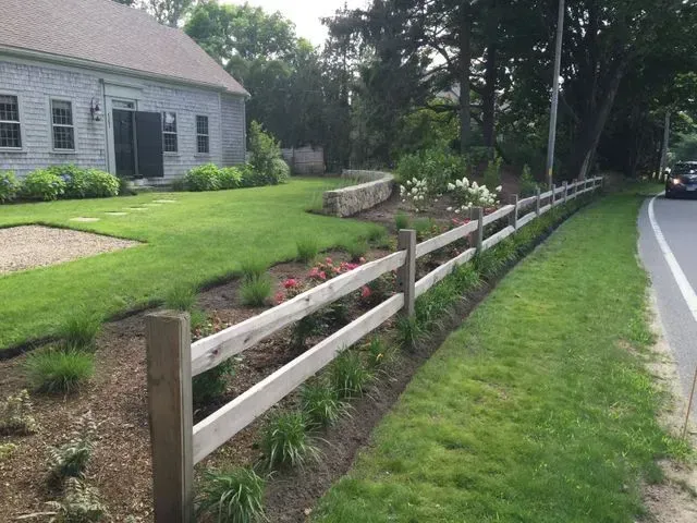 A wooden rail fence borders a landscaped flower bed and lawn in front of a gray house along a roadside.