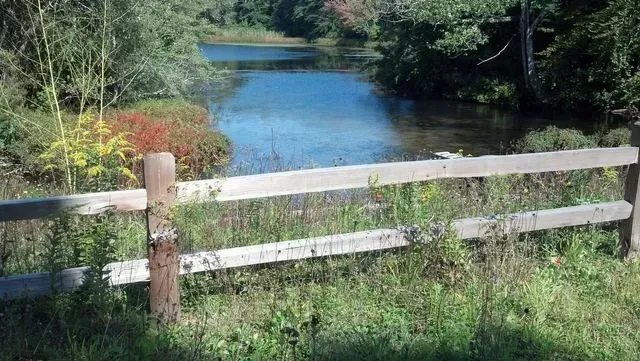 A wooden split-rail fence in the foreground overlooks a calm pond surrounded by trees and colorful autumn shrubs.