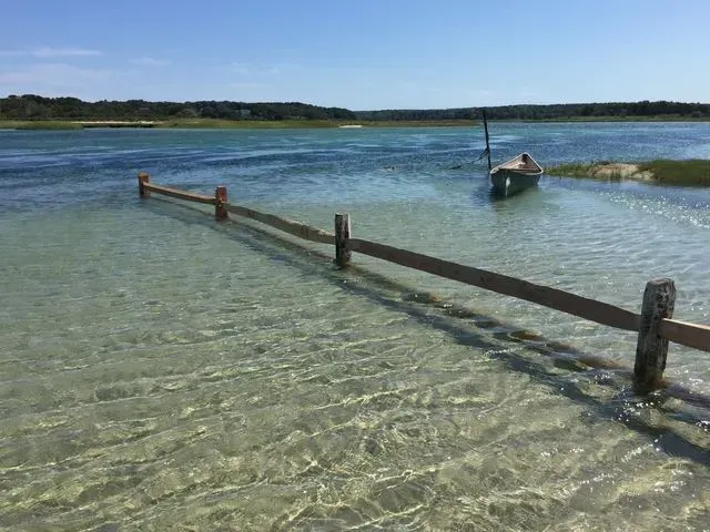 A wooden fence post barrier stands in shallow, clear blue water near a small boat anchored off a grassy shore.