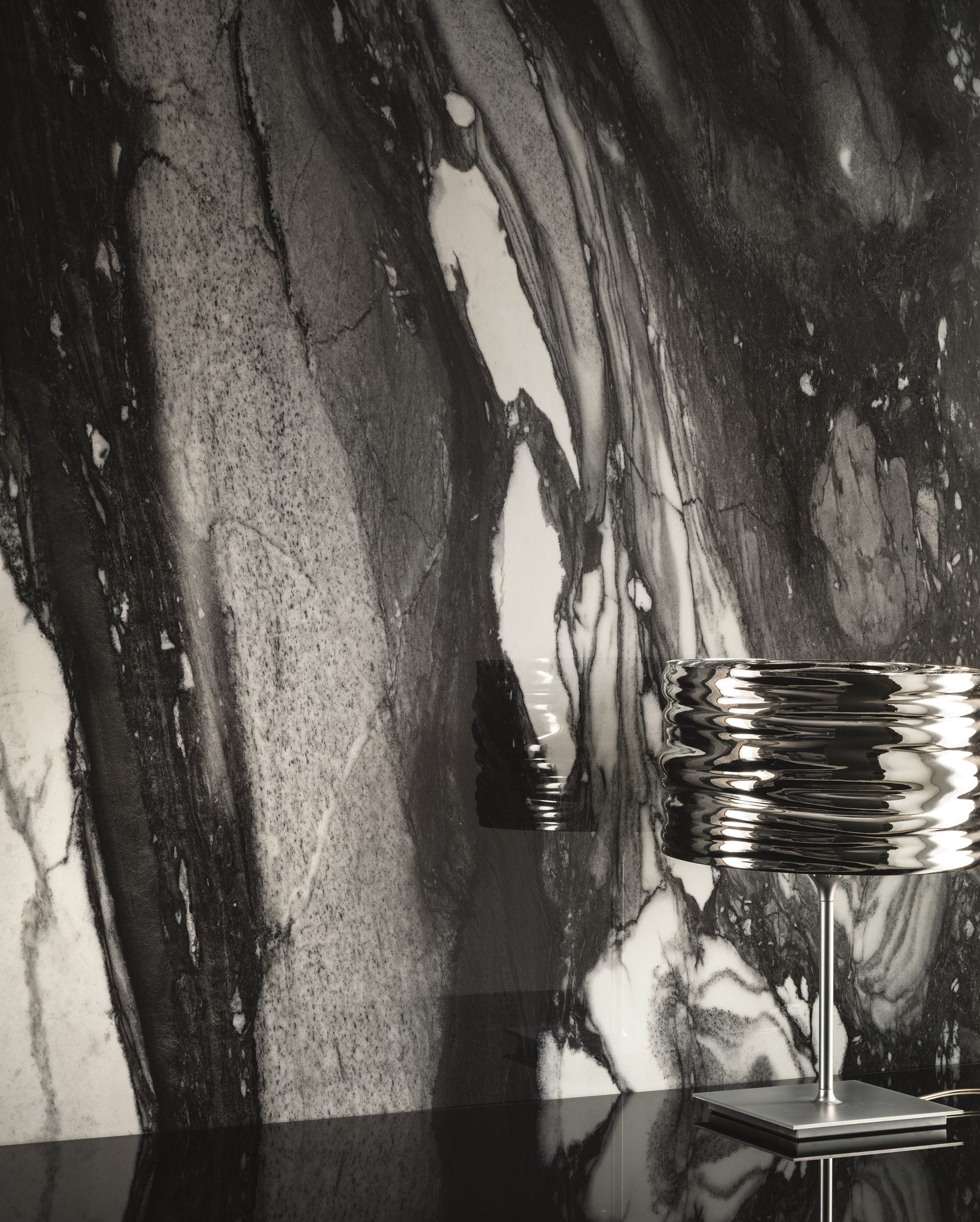 Abstract black-and-white wall with a small metal lamp on a table in the lower right.