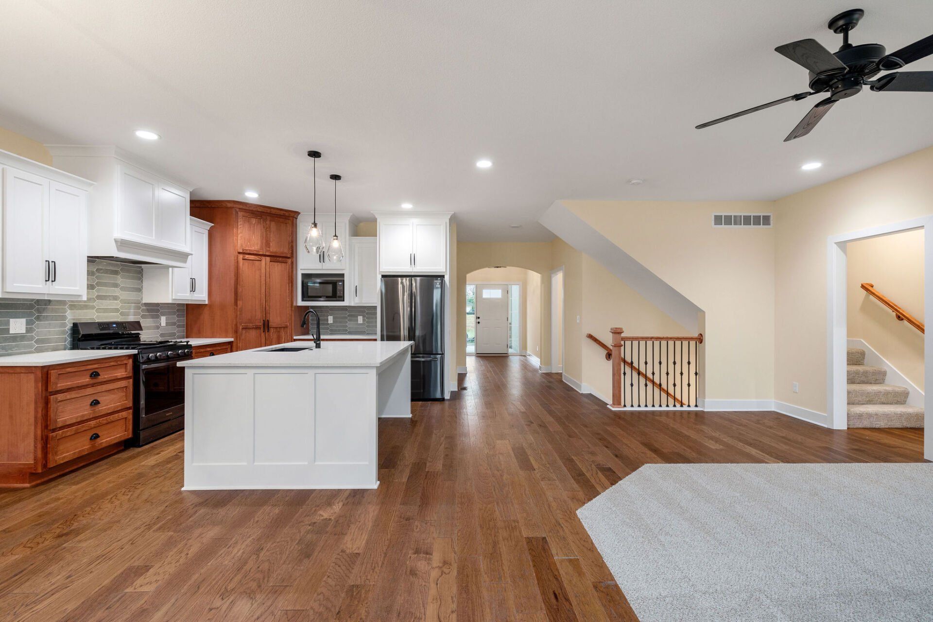 A kitchen with hardwood floors , white cabinets , a refrigerator and a ceiling fan.