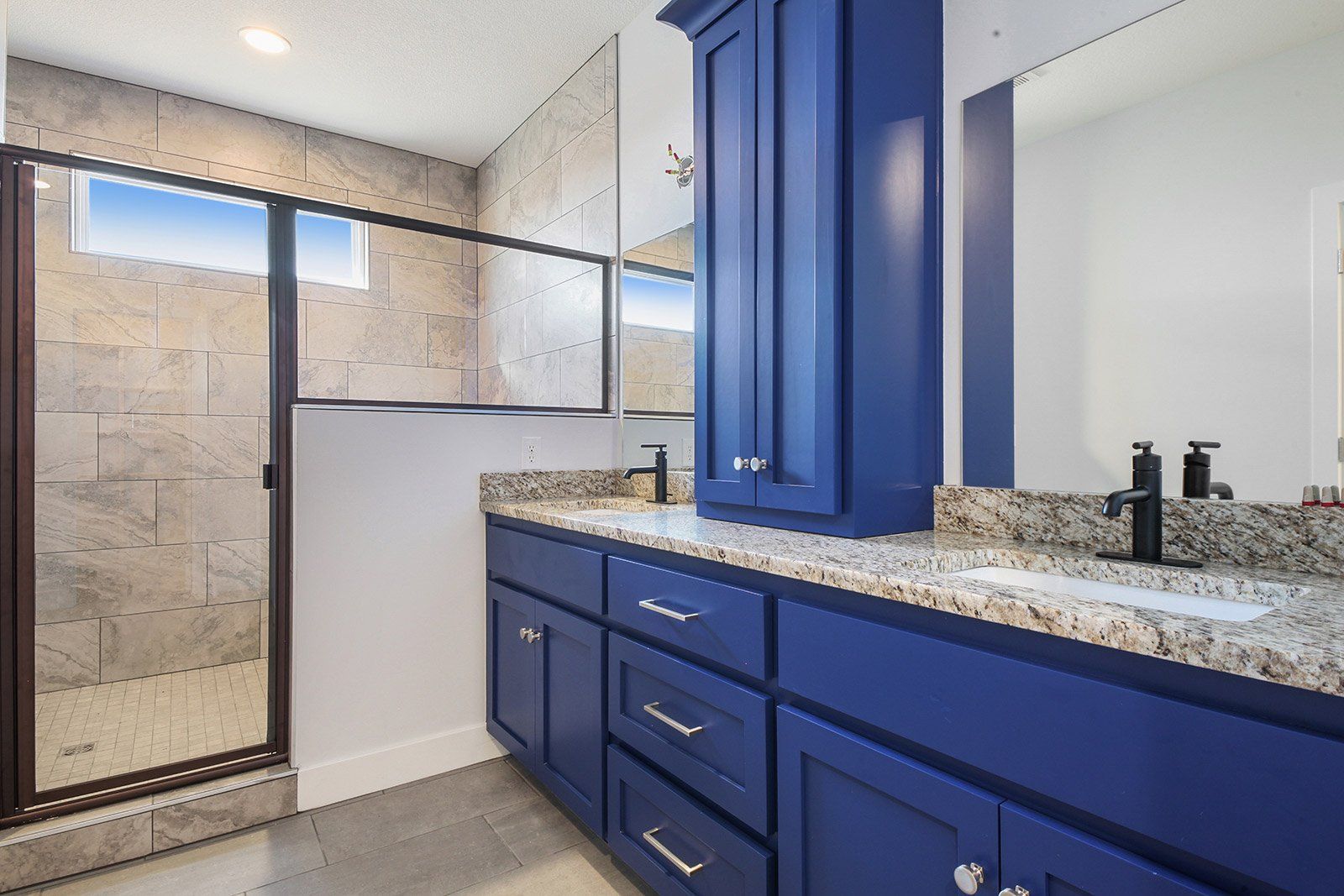 A bathroom with blue cabinets , sinks , mirrors and a walk in shower.