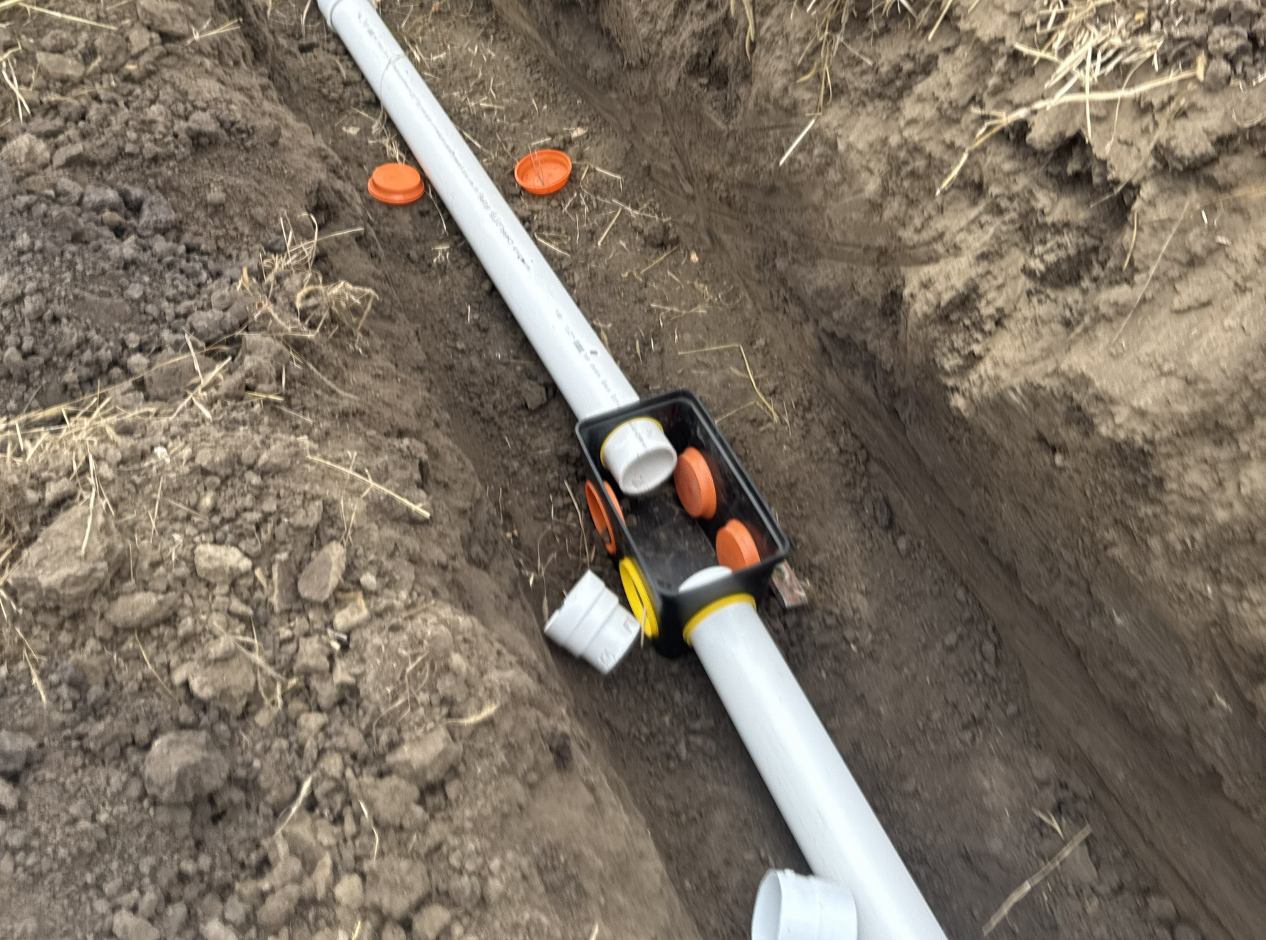 A white PVC pipe and a black irrigation valve box installed in a narrow dirt trench.