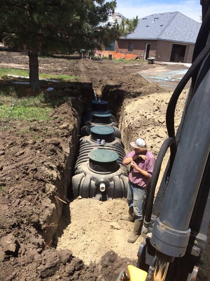 Construction of a septic system: large tanks in a trench, a worker nearby, excavator arm, sunny outdoor setting.