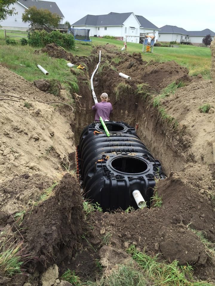 Construction worker installing a large black septic tank in a trench. Pipes, dirt, and houses are in the background.