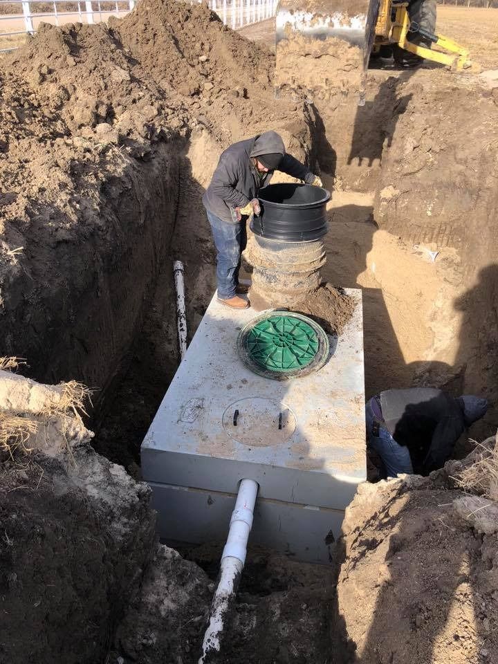 Man installing a septic tank in a trench. Concrete tank, black access lids, and white PVC pipes are visible.