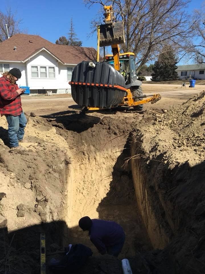 Construction workers installing a septic tank in a trench with a backhoe on a sunny day.