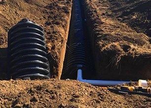 Black corrugated pipes in a trench for drainage; a tank and white pipe are also visible.