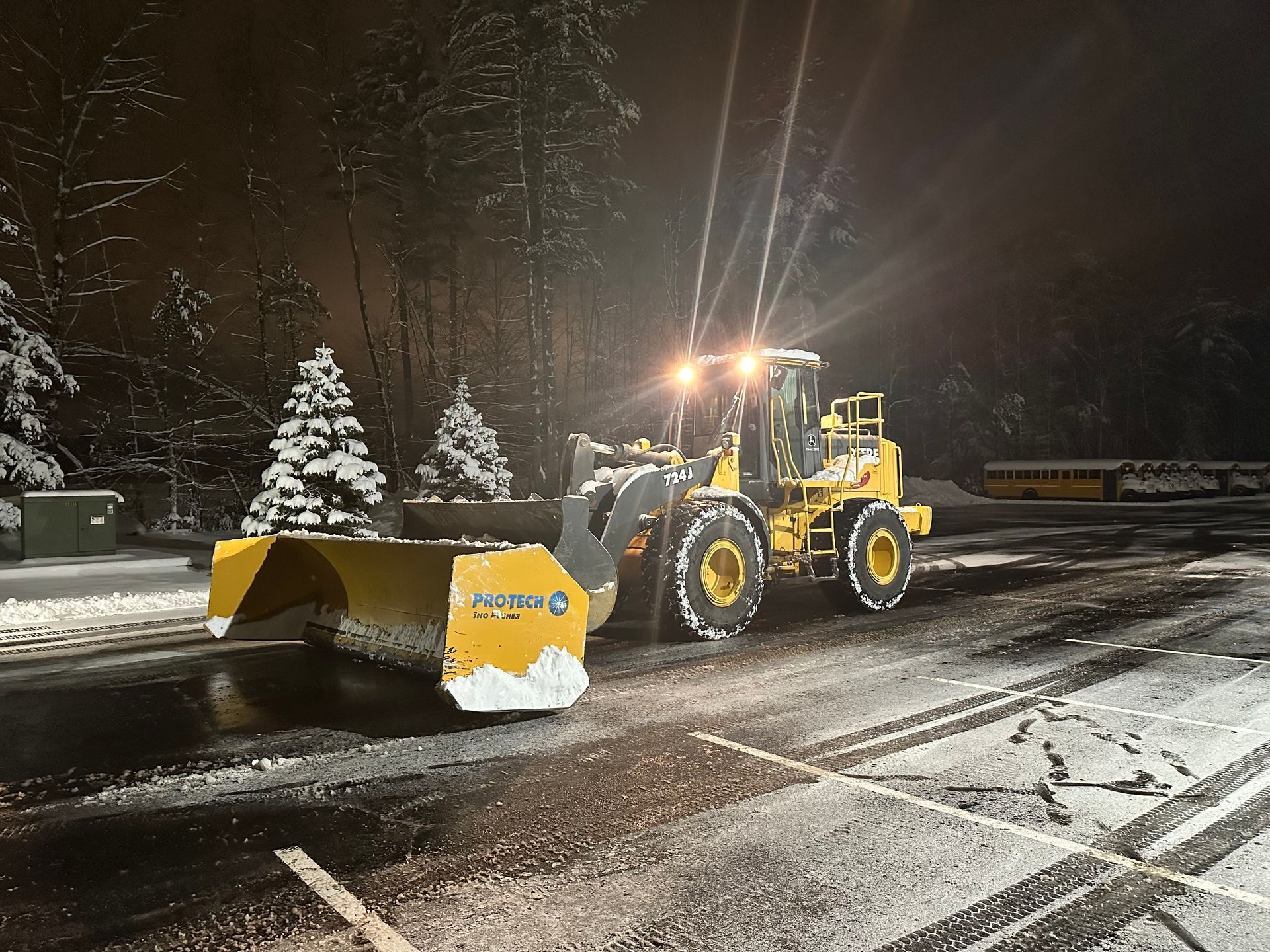 A yellow snow plow is plowing snow in a parking lot at night