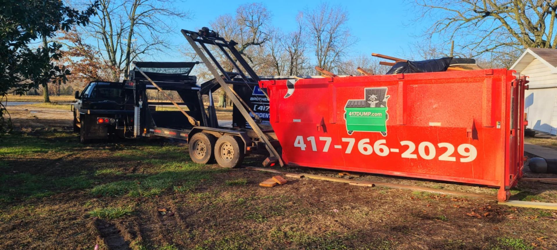 A red dumpster is being lifted by a truck on a trailer, next to a house with trees.
