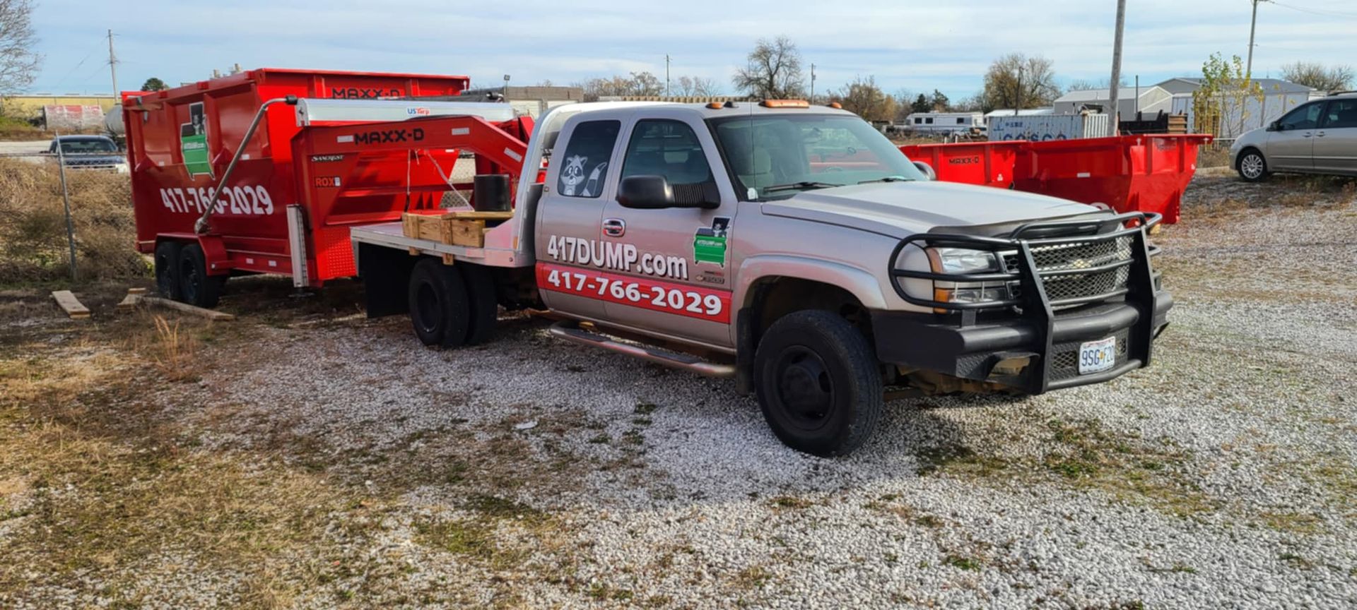A silver pickup truck towing a red dumpster on a gravel lot under a cloudy sky.