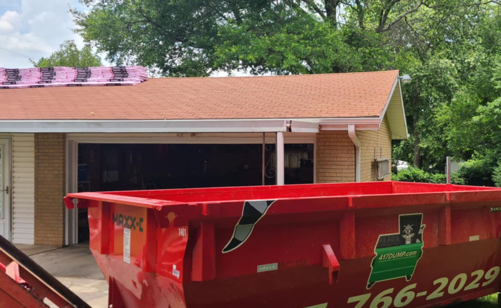 Red dumpster in front of a house with a damaged roof and garage door open.