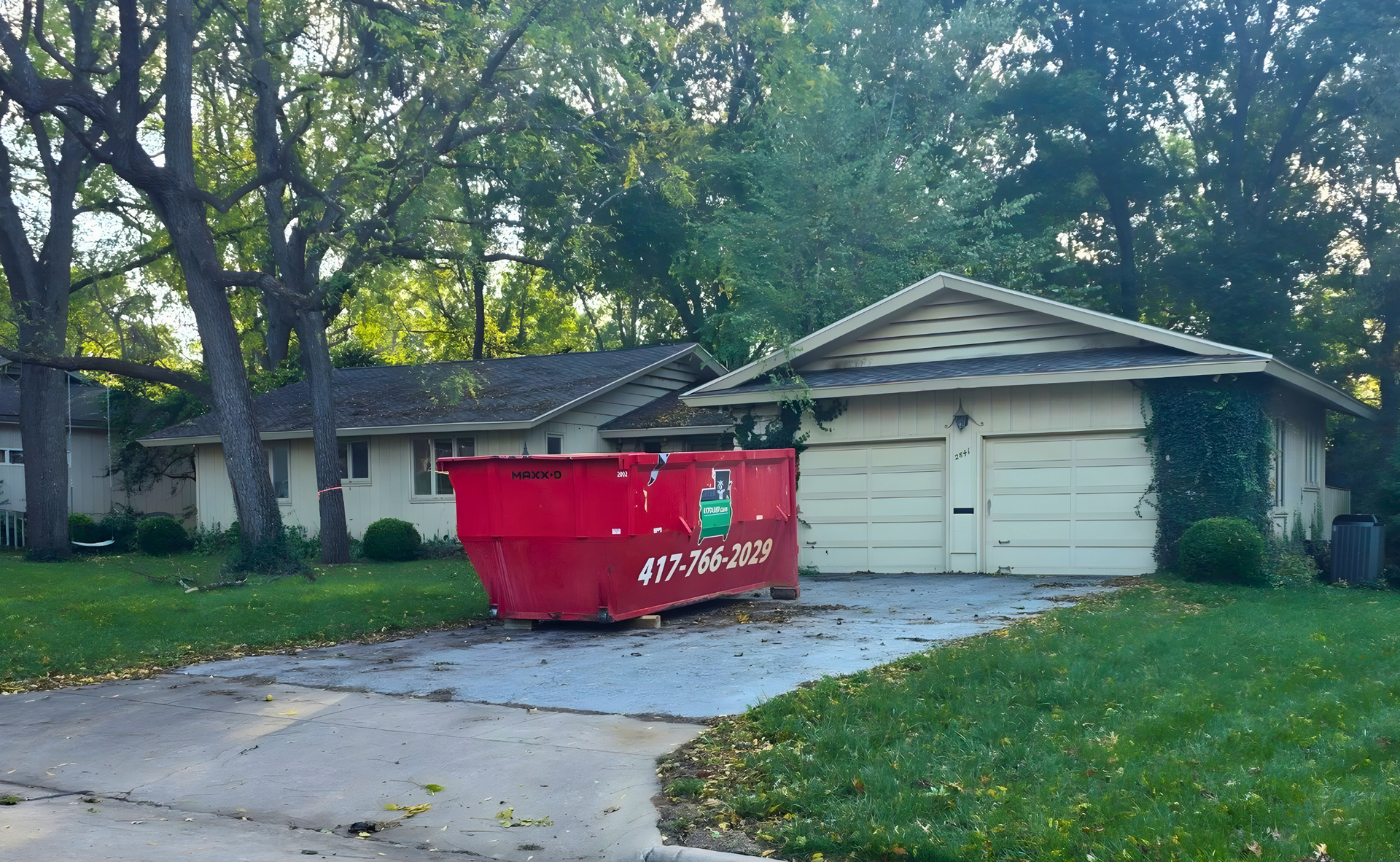 Red dumpster in a driveway beside a beige house and garage. Green lawn and trees surround.