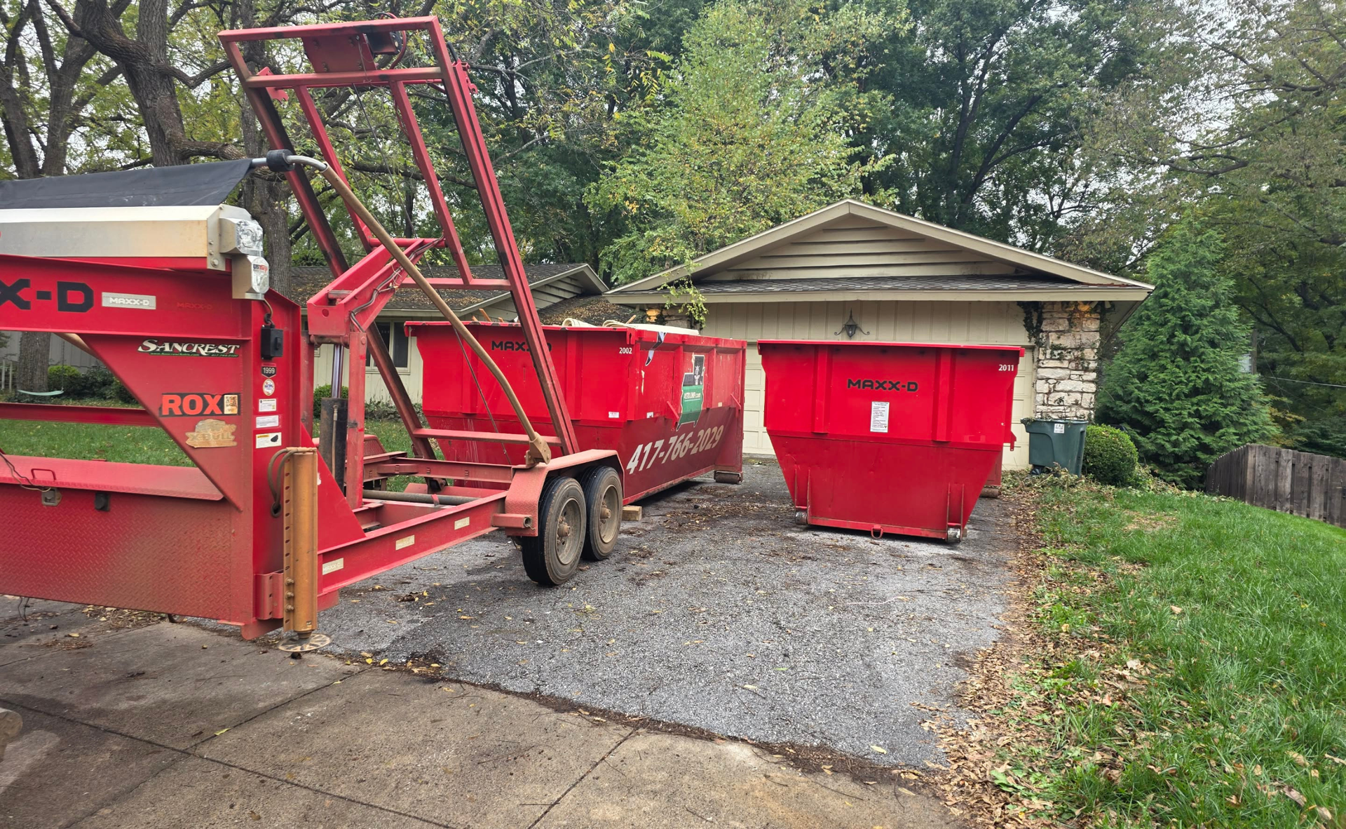 Red dumpster trailer and container on a gravel driveway next to a small house with green lawn.