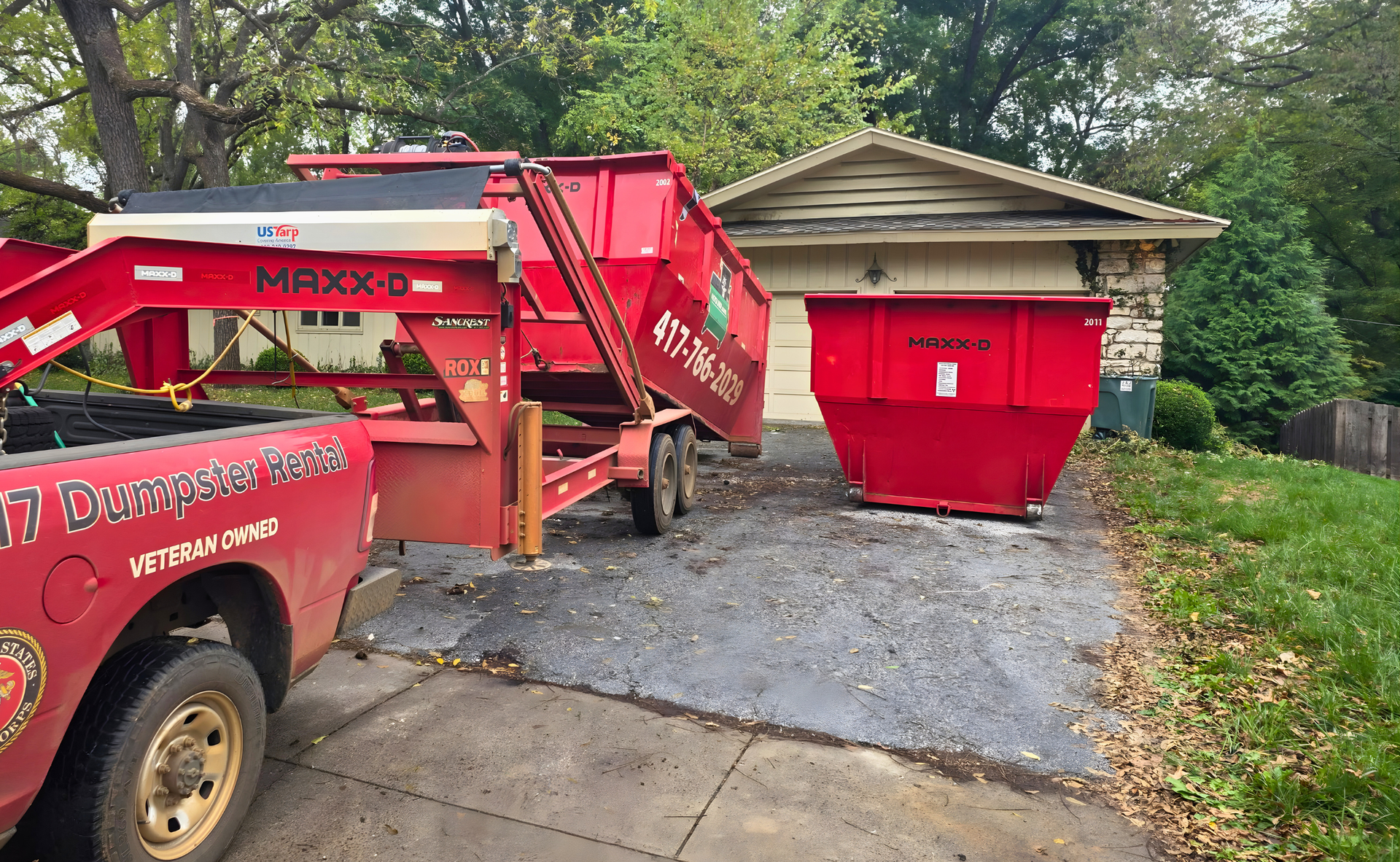 Red dumpster being loaded onto a red truck trailer on a residential driveway next to a garage.