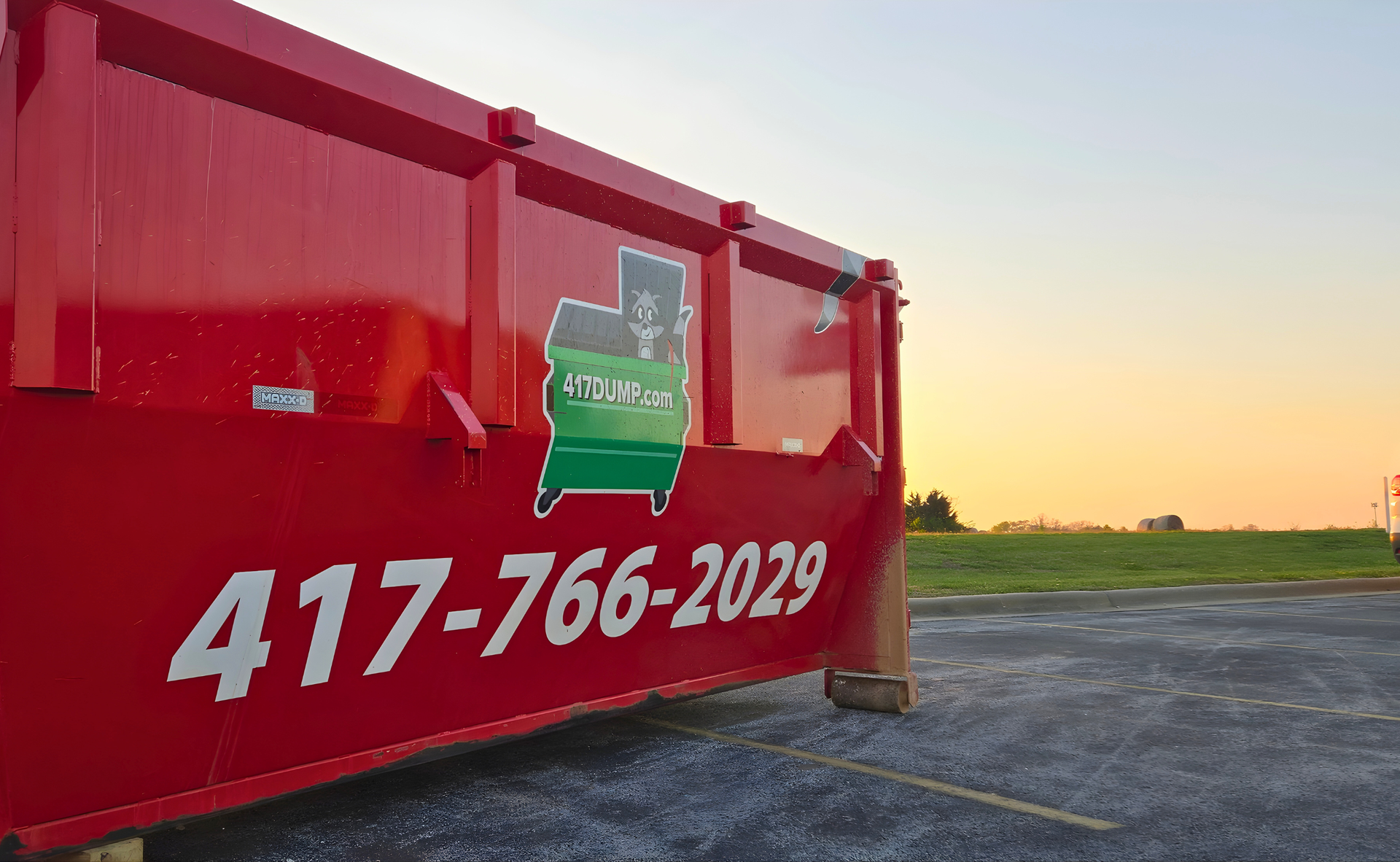 Red dumpster with phone number and logo in parking lot at sunset.