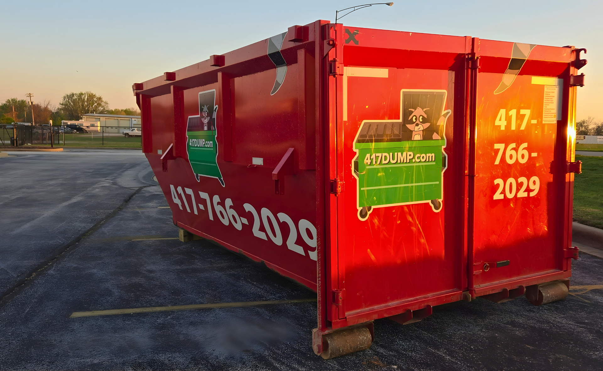 Red dumpster in parking lot with 417-766-2029 phone number and green logo, sunset.