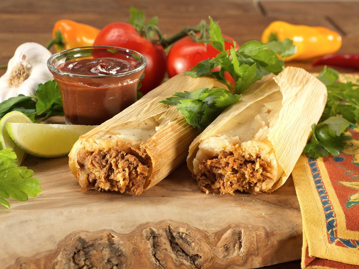 Two tamales on a cutting board with salsa, limes, tomatoes, and cilantro nearby.