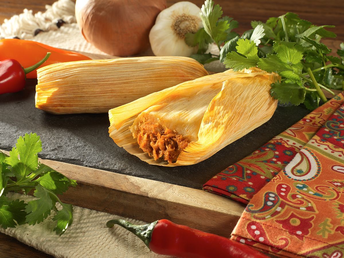 Tamales on a cutting board with chili peppers, cilantro, onions, and patterned cloth