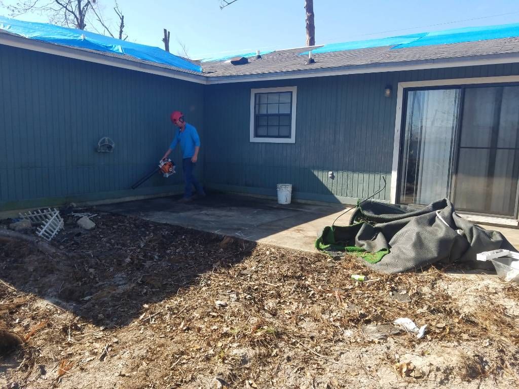 A man is blowing leaves in front of a house with a blue tarp on the roof.