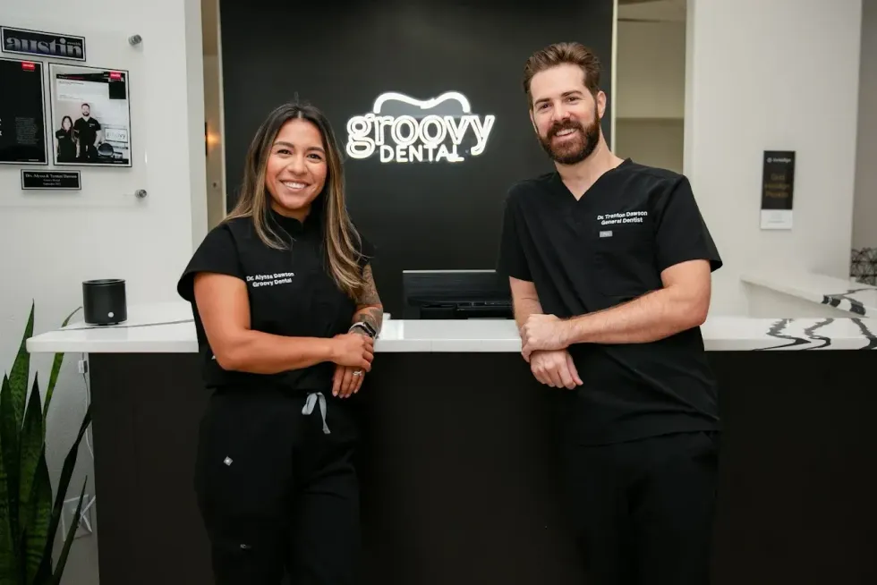 Two people in black uniforms stand behind a counter in a bright office with a “groovy den” sign.