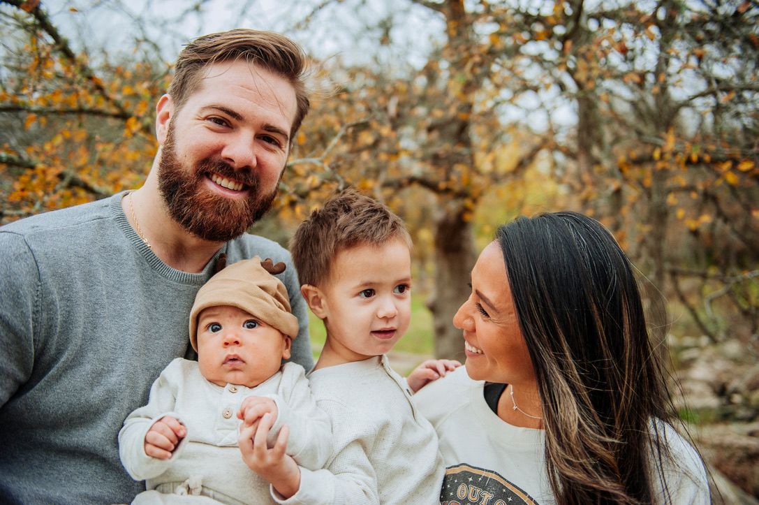 Family of four outdoors in autumn, smiling and holding a baby.