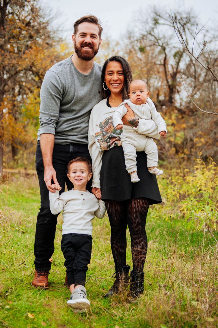 Family of four standing together outdoors in a grassy field with autumn trees in the background.