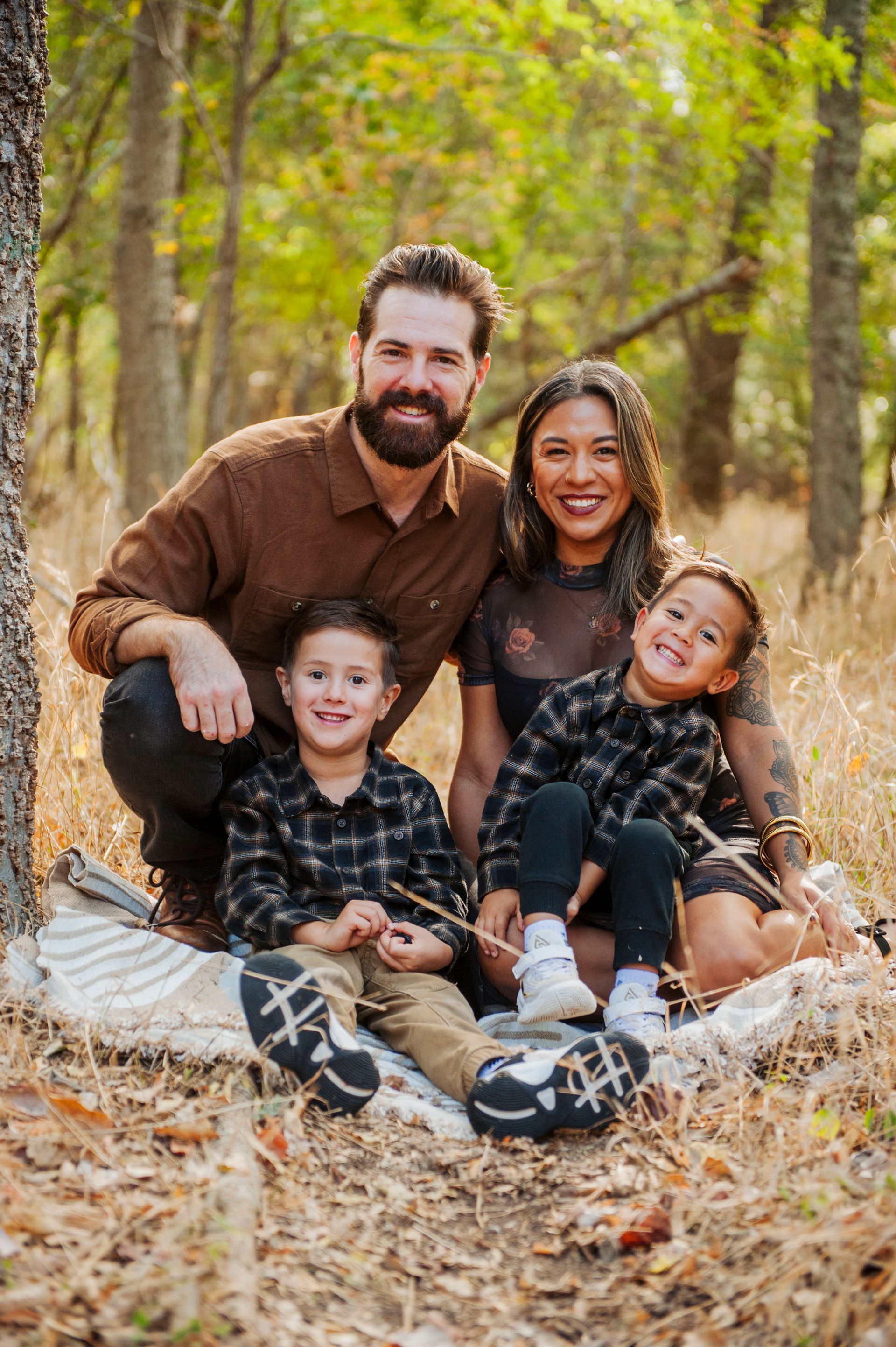 Family of four sitting on a leaf-covered path in a sunlit wooded area, smiling at the camera